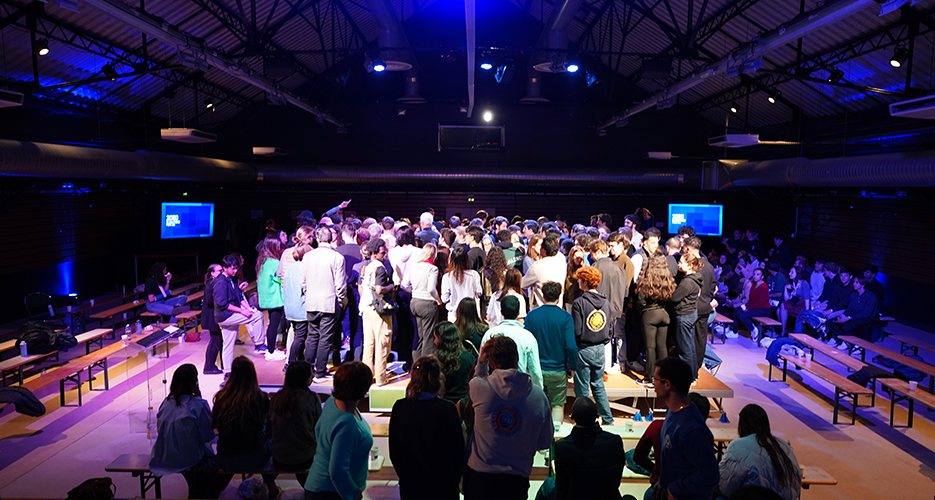 Large group of people gathered in the center of an indoor event space with industrial-style ceilings and multiple screens displaying text, surrounded by seated audience members.