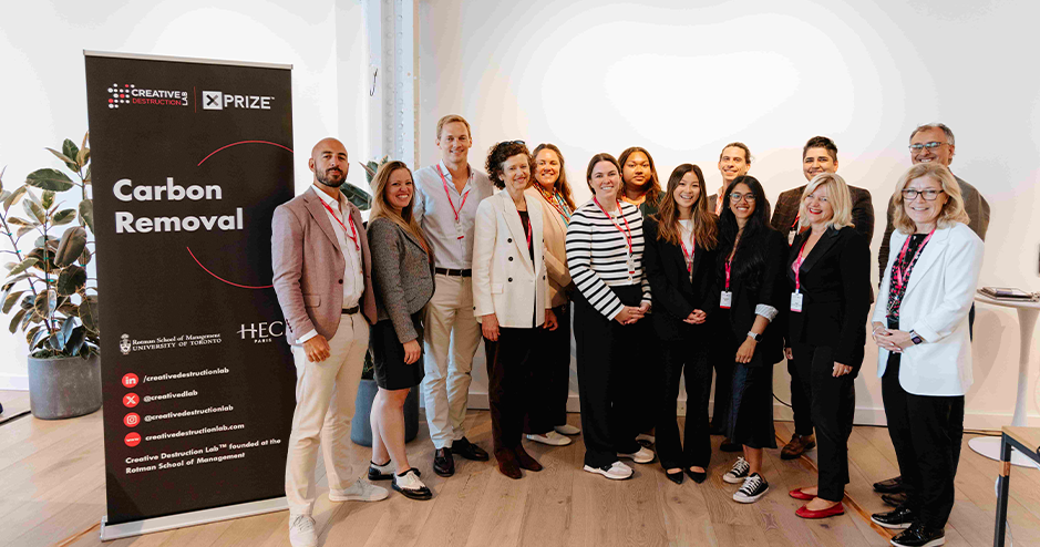 Group photo of thirteen people standing next to a 'Carbon Removal' banner at a Creative Destruction Lab XPRIZE event. The individuals are dressed in business attire with name badges, smiling, and posed in a brightly lit room with white walls and wooden floors.