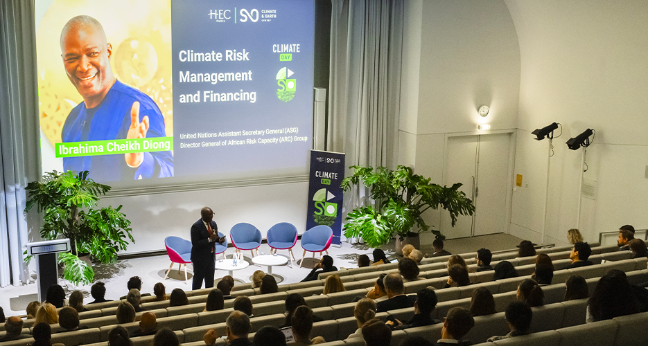 A speaker addresses an audience in a lecture hall during the 'Climate Day' event at HEC Paris. The projection screen behind him displays a slide with the title 'Climate Risk Management and Financing' and a photo of Ibrahima Cheikh Diong