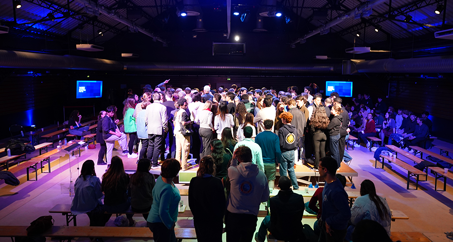 A large group of people gathered in the center of an indoor event space, illuminated by blue and purple lighting. The setting includes industrial-style ceilings with exposed beams and spotlights. The audience is standing closely together, likely engaging in a group activity or discussion