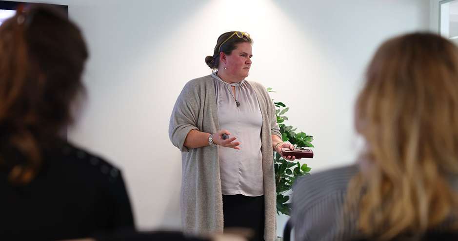 Marcelle Laliberté giving a presentation in a softly lit room. She is dressed in a light cardigan and blouse, holding a small device in one hand and gesturing with the other