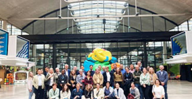 Group of executives and professionals gathered at a modern innovation hub, with a colorful sculpture and industrial-style architecture in the background