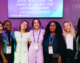 seven smiling women pose together at the Women’s Forum event. They are dressed in professional attire and wear accreditation badges around their necks. Behind them, a purple backdrop features a large screen with the text 'GOOD: Reweaving the Fabric of Society, One Venture at a Time Pitching Contest.' The women are standing close to each other, conveying a sense of camaraderie and positivity