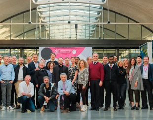 Photo de groupe d'un ensemble de professionnels posant dans un espace moderne avec une verrière en arrière-plan. Une bannière rose au centre affiche le texte 'CREATIVE DESTRUCTIONS LAB'. Les participants portent des tenues professionnelles et certains ont des badges roses autour du cou, indiquant un événement ou une conférence.