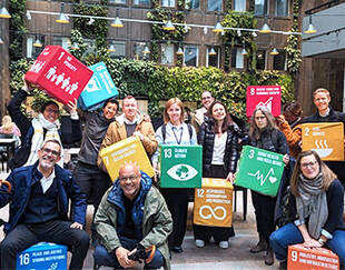 A diverse group of people posing in an indoor space with green walls, holding colorful blocks representing the United Nations Sustainable Development Goals (SDGs)