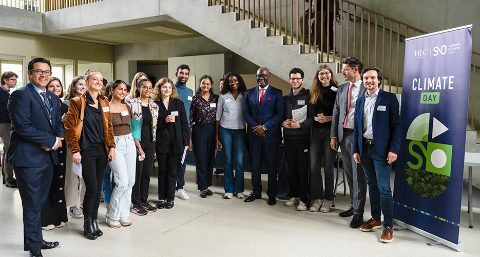 A group of people, including students and professionals, posing together at the 'Climate Day' event at HEC Paris. The image shows smiling participants, some holding certificates or documents, standing in front of a concrete staircase inside a modern building. On the right side of the image, a banner displays the logos of HEC Paris and Climate and Earth, with the text 'Climate Day'