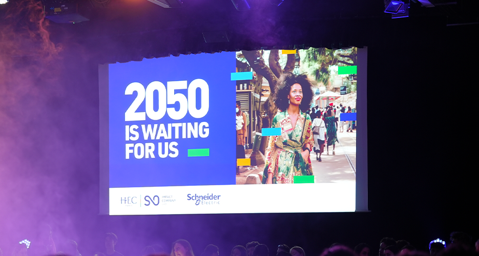 A large indoor event with a stage and an audience seated in a dimly lit room. A screen displays the message '2050 IS WAITING FOR US' along with the logos of HEC Paris, SO, and Schneider Electric